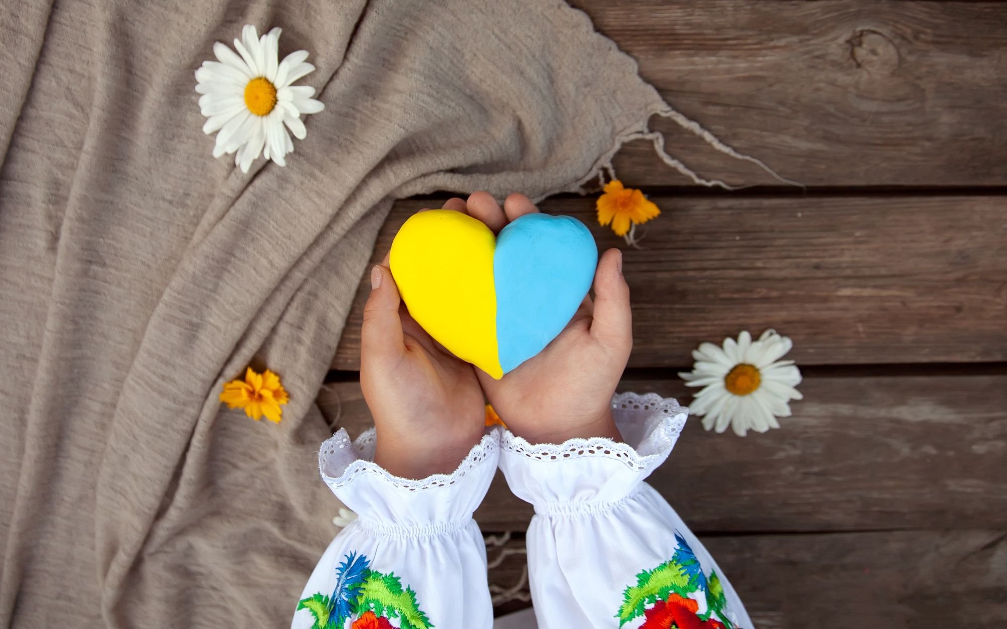 A yellow and blue heart in the hands of a child in an embroidered shirt, against the background of a rough wooden table.
